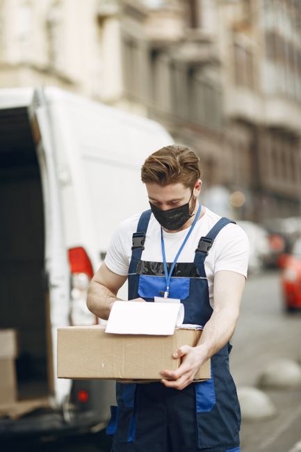 A young man with a beard, wearing a red cap backwards, a multi-tone beige and brown jacket over a blue checked shirt, and striped gloves, sits on the edge of a moving van with the rear door open. Behi