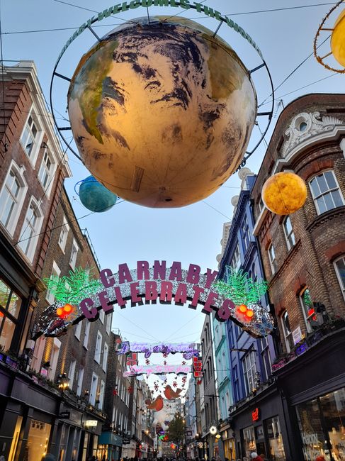 A large, colorful sign spelling 'CARNABY' in individual letters with vibrant, multicoloured lighting hangs above a narrow street, marking the entrance to Carnaby Street in London. Below the sign, a se